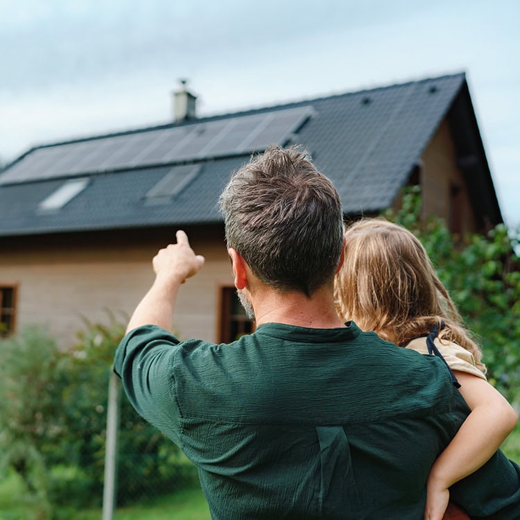 En pappa håller sitt barn i famnen och pekar bort mot hustaket som har en solcellspanel installerad. En symbol för framtiden och vikten av klimatvänliga val.